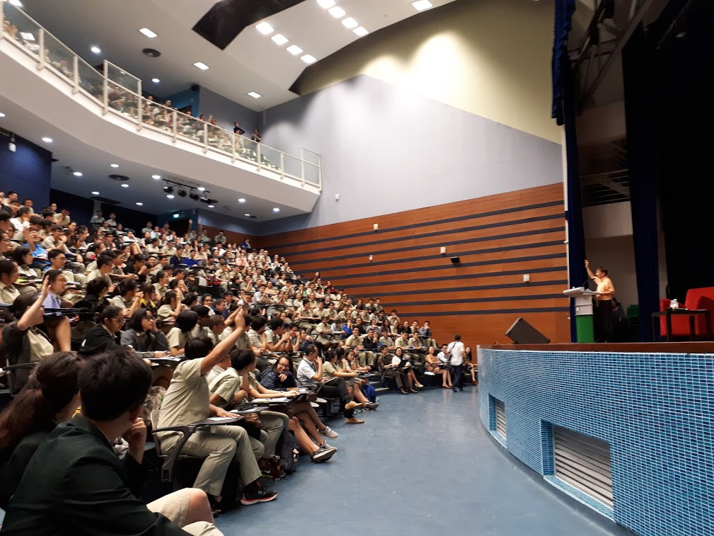 A packed auditorium with students in uniform seated in tiered rows facing a speaker at a podium on stage.